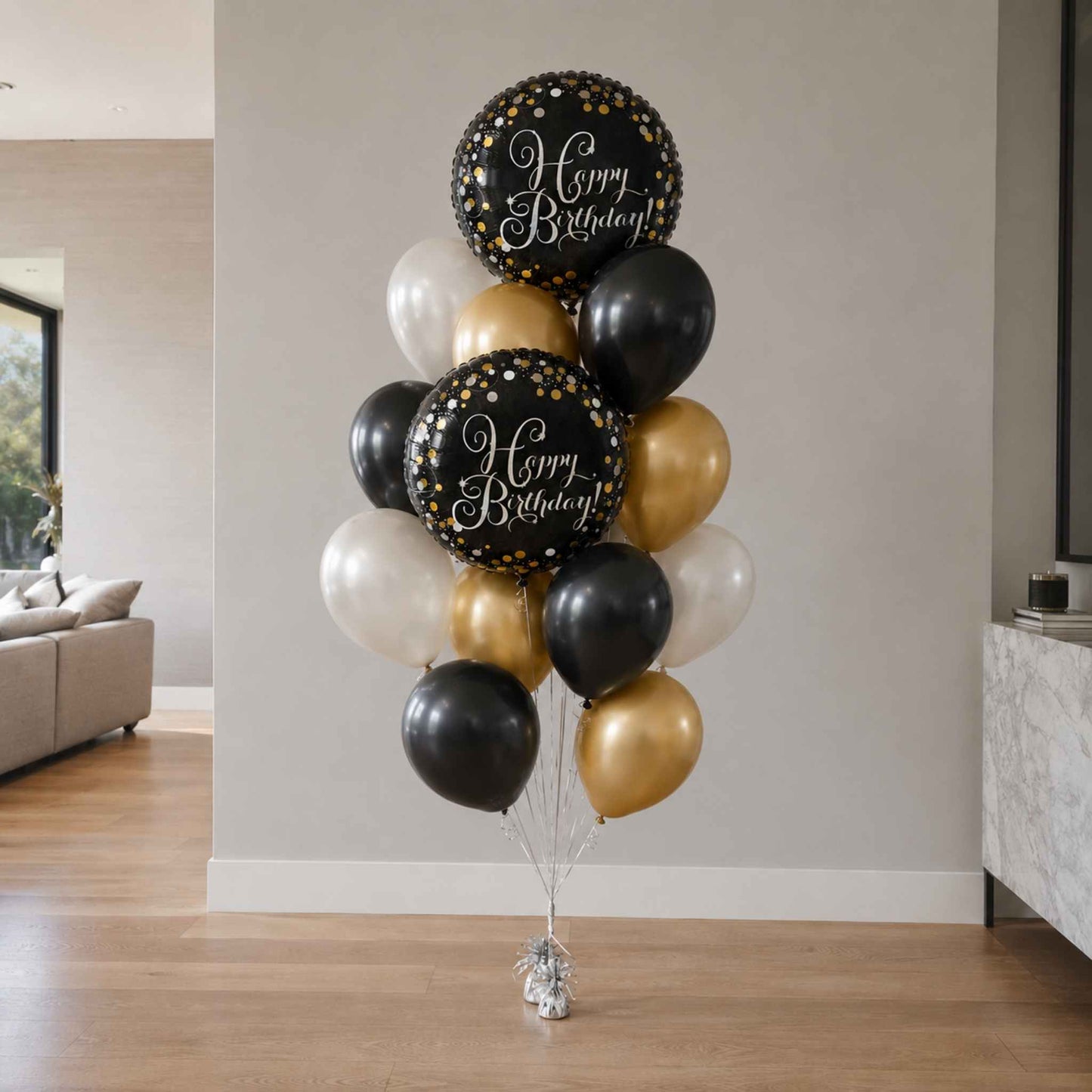 A balloon bouquet cluster with two large black balloons printed with 'Happy Birthday!' in white script, surrounded by black, white, and metallic gold balloons, displayed indoors against a light gray wall.