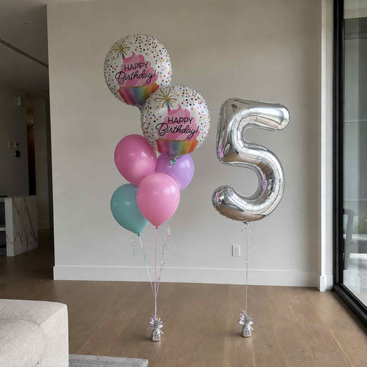 A birthday balloon bouquet and a large metallic number 5 balloon in a modern indoor room with wood floors and a light wall.