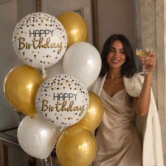 A woman in a champagne-colored dress holds a glass of white wine beside a cluster of metallic gold and white balloons printed with 'HAPPY Birthday' and gold confetti, in an indoor setting.