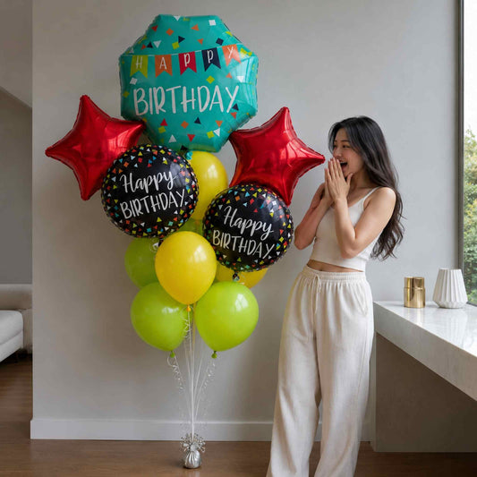 A woman in a white crop top and cream pants stands beside a cluster of birthday balloons in a bright indoor room. The balloon set includes a teal geometric balloon reading 'HAPPY BIRTHDAY,' two red star balloons, two black round balloons with confetti and 'Happy Birthday' text, and several yellow and lime-green balloons.