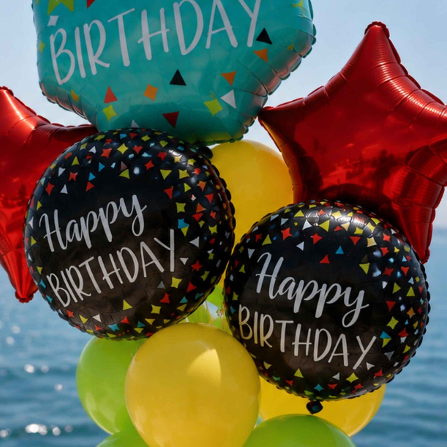 A cluster of birthday balloons including two round black balloons with 'Happy Birthday' text and multicolor confetti, a teal foil balloon, a red star foil balloon, and yellow balloons, set against a blue sky and water background.