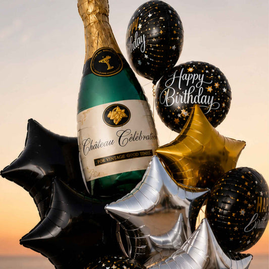 A celebratory balloon bouquet featuring a green champagne bottle, black and gold balloons, and metallic star-shaped balloons in black, gold, and silver, set against a warm sunset sky.