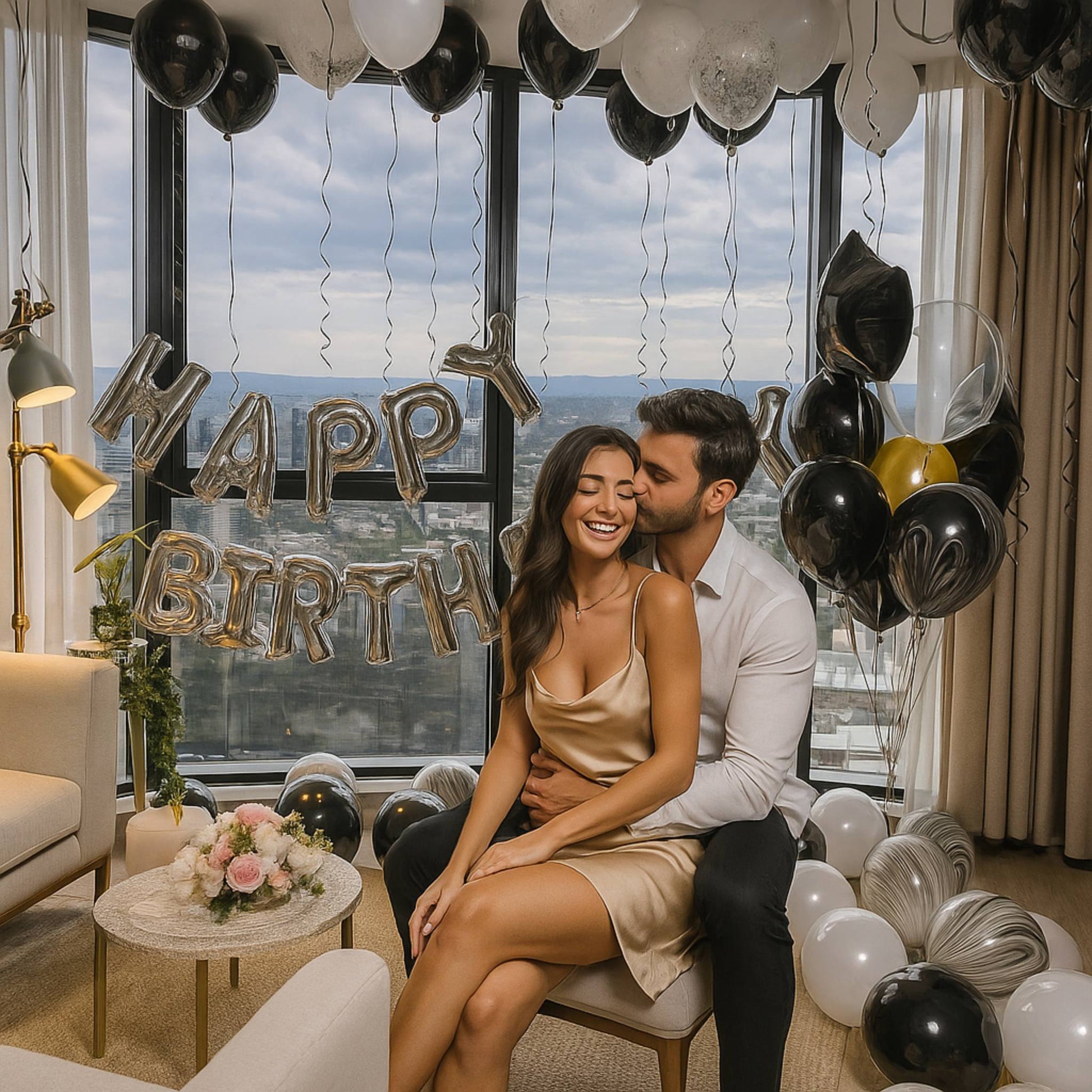 Couple celebrating a birthday with balloons and a 'Happy Birthday' sign in a room with large windows.