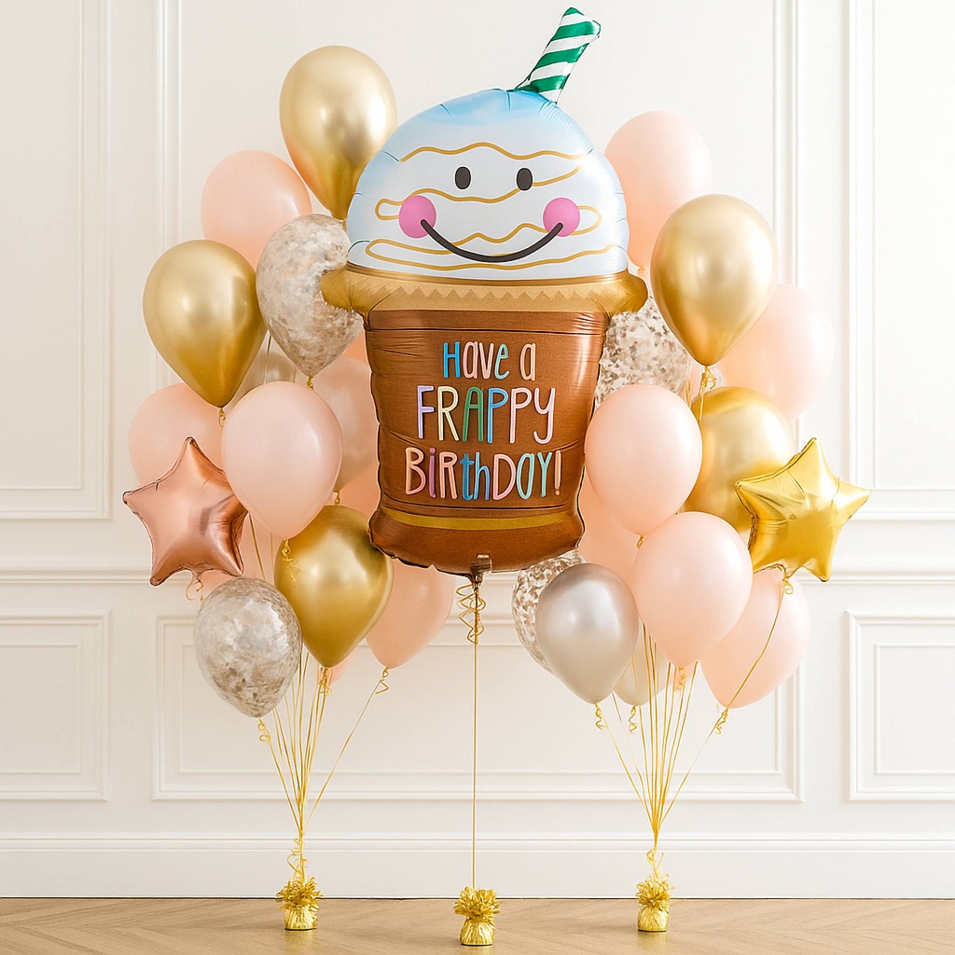 Balloon bouquet with a cake-shaped balloon and 'Happy Birthday' text, surrounded by gold, pink, and star-shaped balloons on a white wall background.