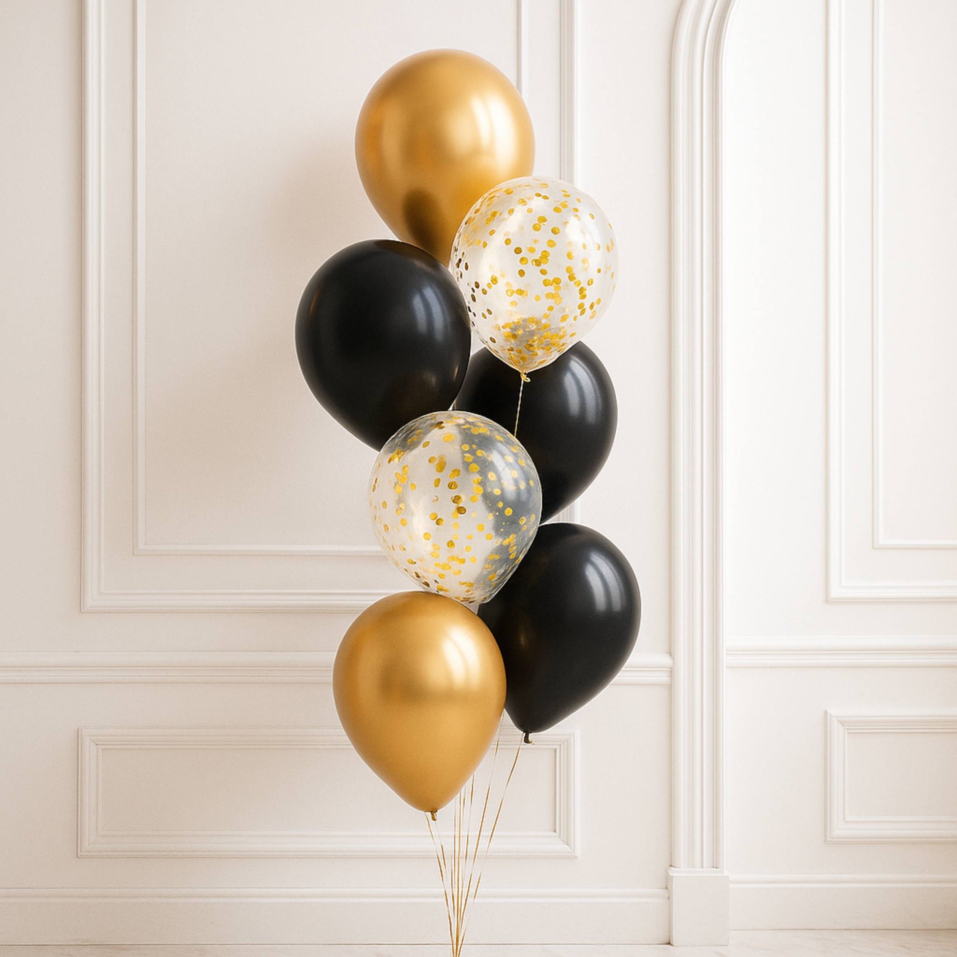 Bouquet of gold, black, and clear confetti balloons against a white wall.