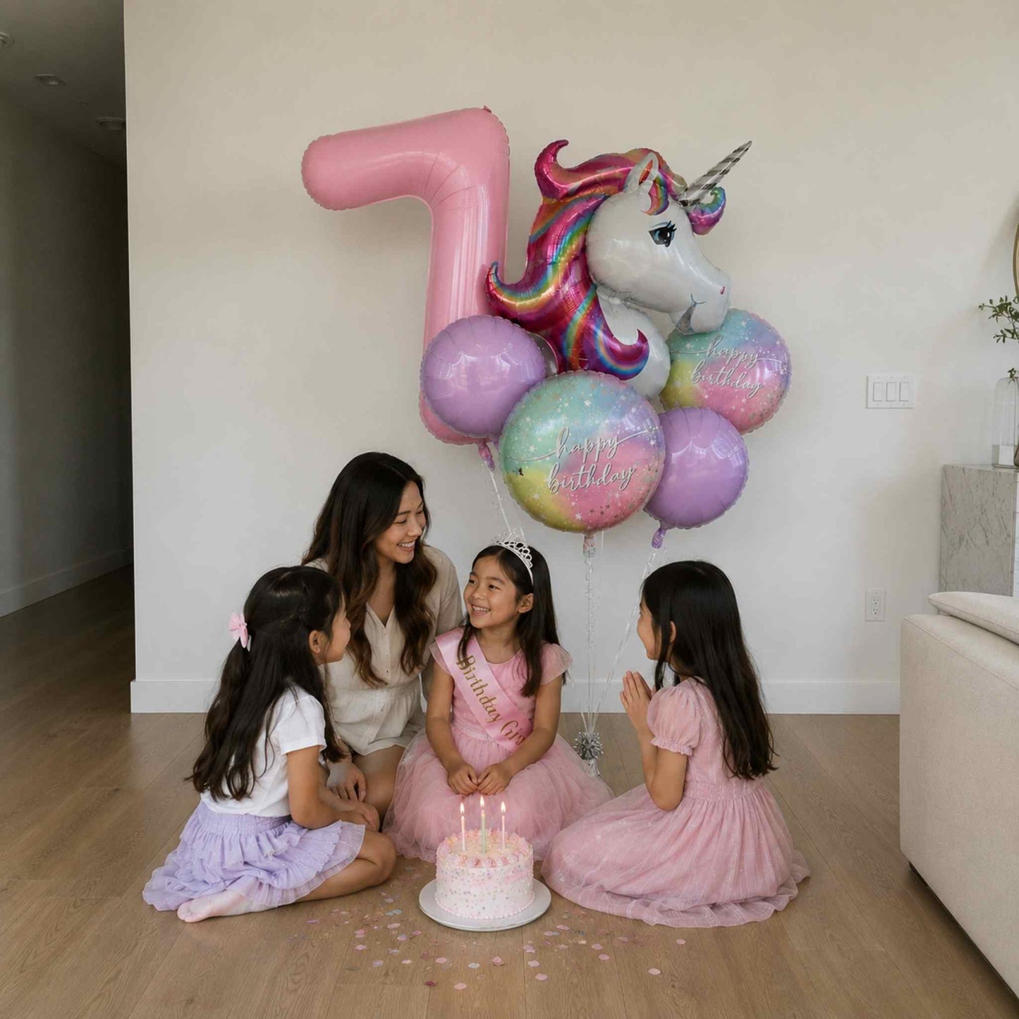 A woman and three young girls sit on a wooden floor in a bright room, gathered around a small birthday cake with candles. Large decorative balloons, including a pink number 7 balloon and a unicorn head balloon, float behind them.