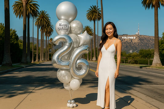 Woman in a white dress standing next to large silver balloons with 'Happy Birthday' and number '24' in front of the Hollywood sign.