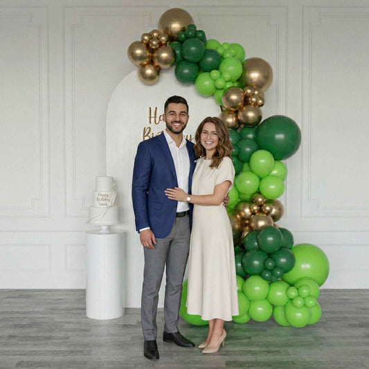 Man and woman posing in front of a balloon arch with green and gold balloons.