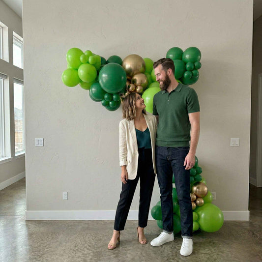 Man and woman standing in a room with a large heart-shaped arrangement of green and gold balloons.