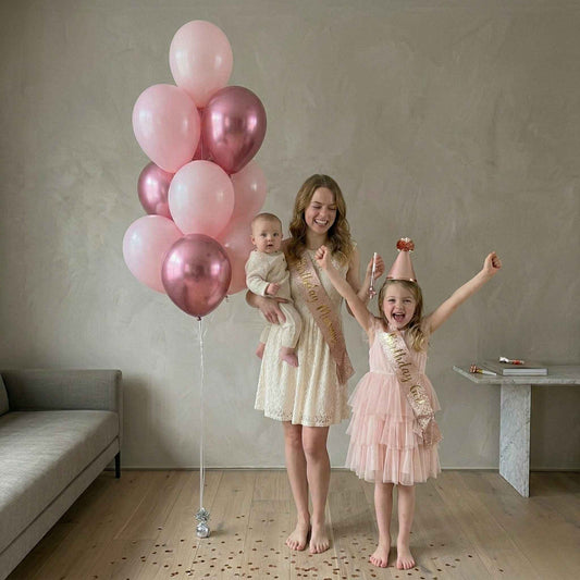 Woman and two children with pink balloons and sashes in a room.