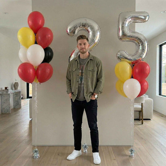 Man standing between two large silver number 25 balloons with colorful balloons on either side in a room.