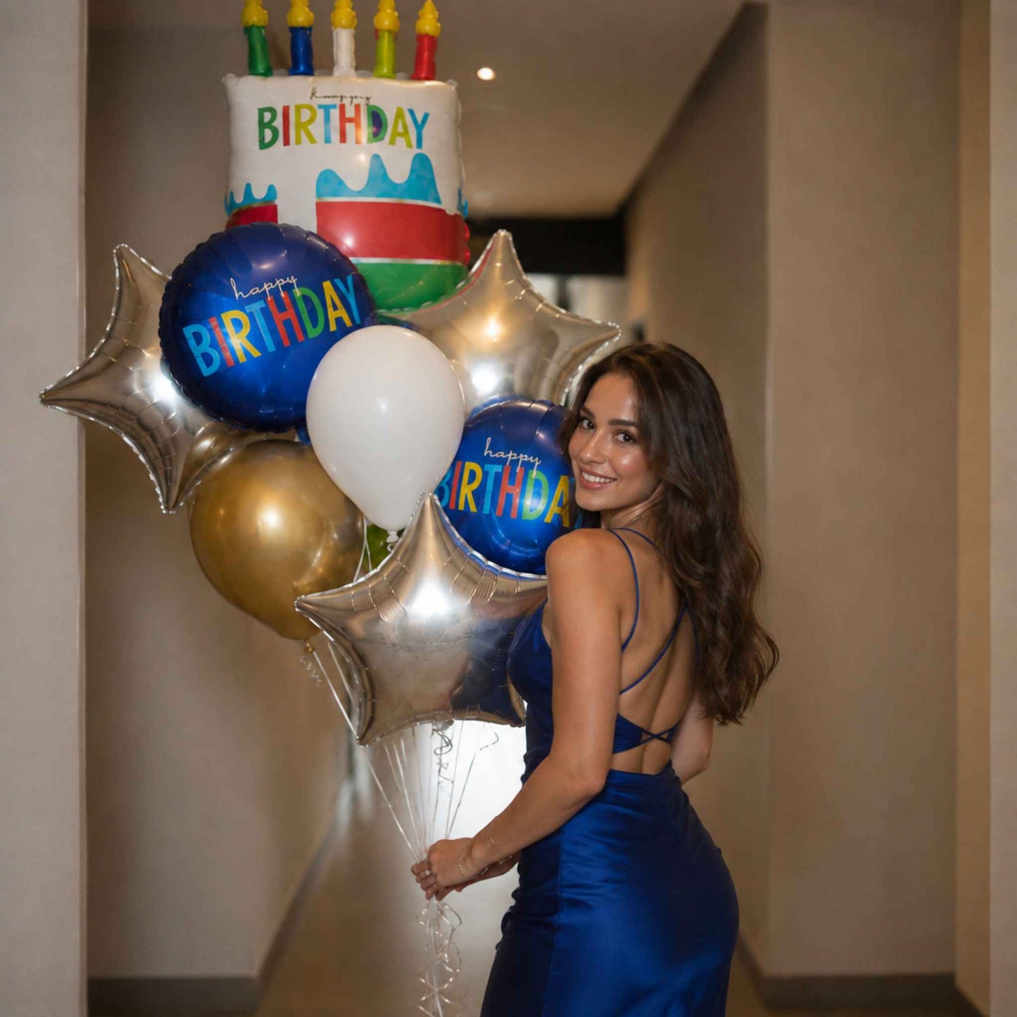 A woman in a blue satin dress holds a birthday balloon bouquet featuring a white cake-shaped balloon, blue and gold round balloons, and silver star balloons in a hallway.