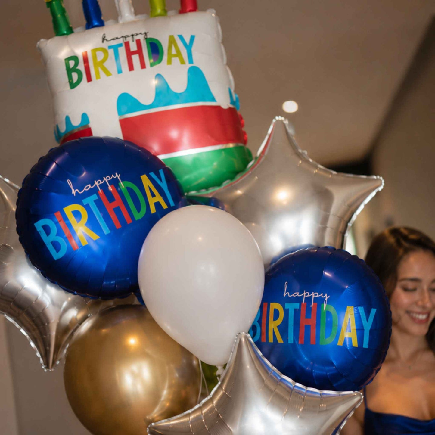 A birthday balloon bouquet featuring a cake-shaped balloon with candles, blue foil balloons reading 'happy birthday,' a white round balloon, metallic silver star balloons, and a gold balloon, with a smiling woman partially visible in the background.