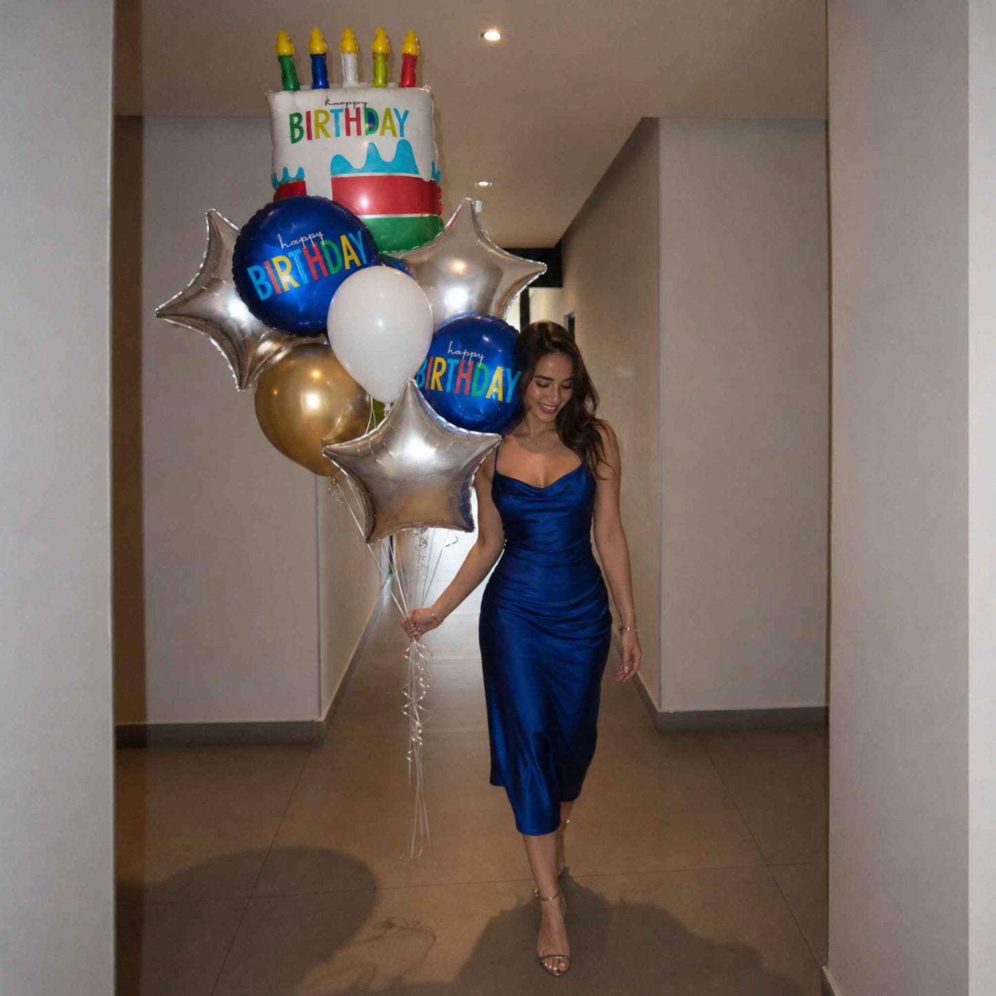 A woman in a shiny royal blue satin midi dress walks down a hallway holding a cluster of birthday balloons, including a cake-shaped balloon, silver star balloons, blue balloons with 'BIRTHDAY' text, and a white balloon.