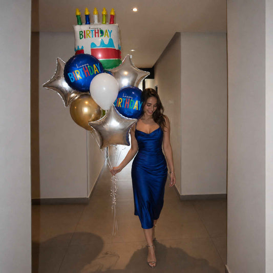A woman in a shiny royal blue satin midi dress walks down a hallway holding a cluster of birthday balloons, including a cake-shaped balloon, silver star balloons, blue balloons with 'BIRTHDAY' text, and a white balloon.