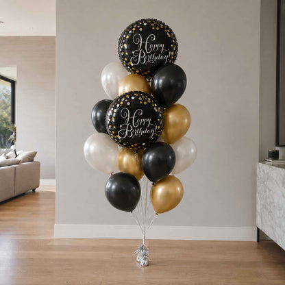A balloon bouquet cluster with two large black balloons printed with 'Happy Birthday!' in white script, surrounded by black, white, and metallic gold balloons, displayed indoors against a light gray wall.