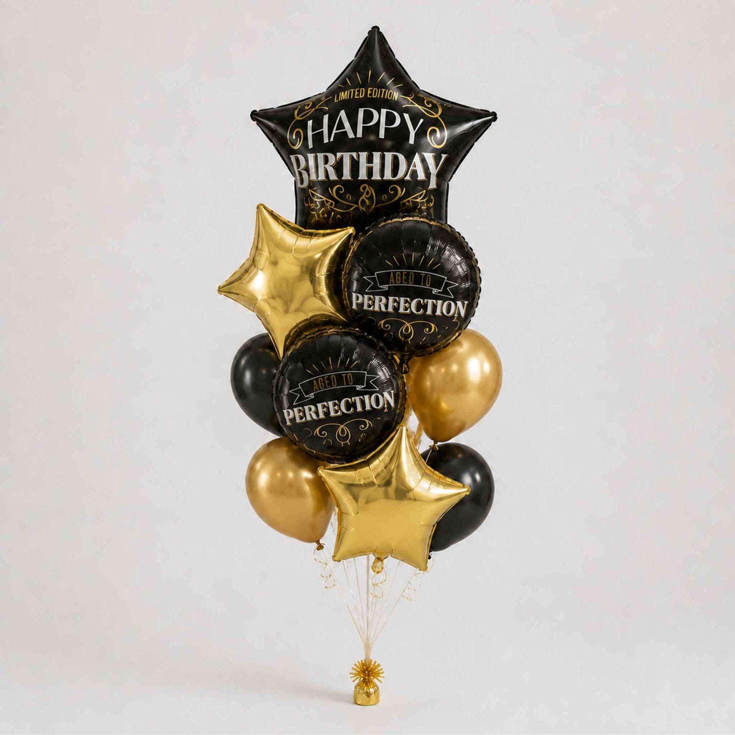 A balloon bouquet featuring a large black star-shaped foil balloon with gold 'HAPPY BIRTHDAY' lettering, two black round balloons with gold 'PERFECTION' text, two metallic gold star-shaped balloons, and two metallic gold round balloons, arranged against a light gray studio background.