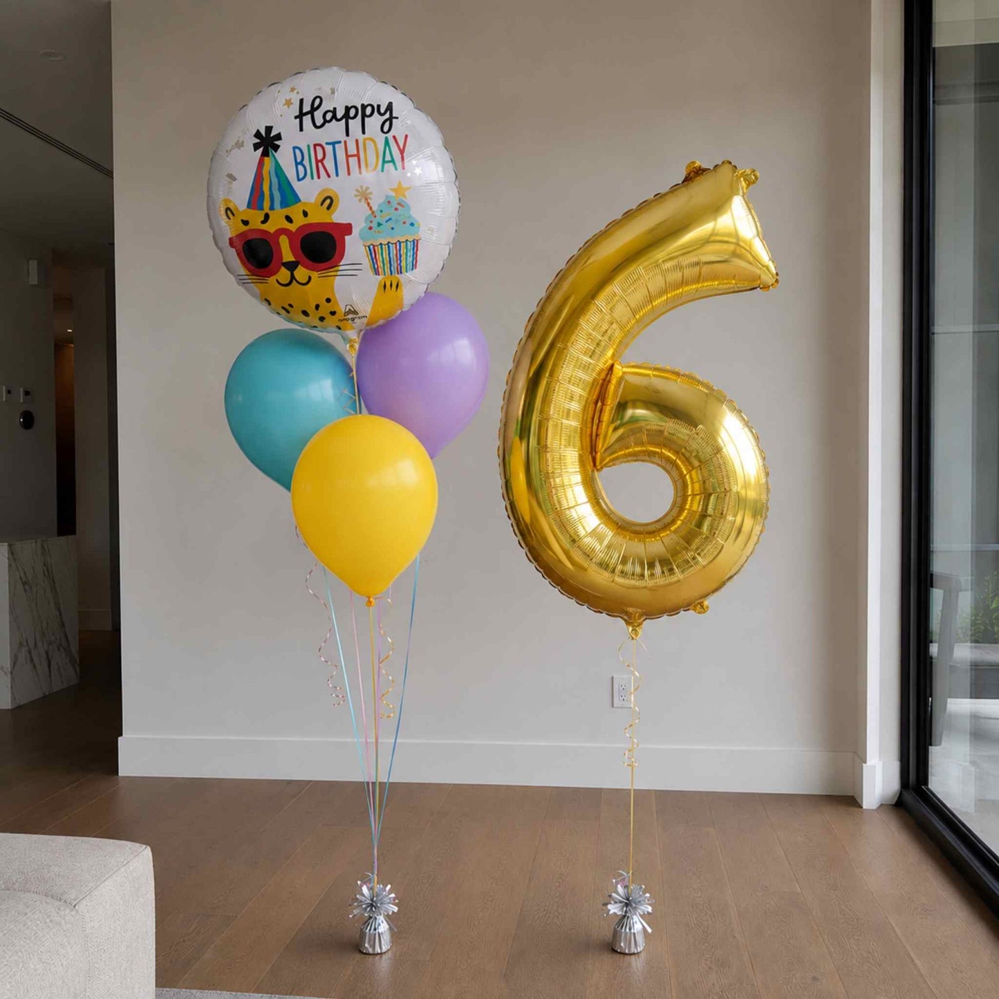 A set of birthday balloons in a room, including a large metallic gold number 6 foil balloon, a round white 'Happy Birthday' balloon with a cartoon leopard, and three smaller balloons in teal, yellow, and purple.