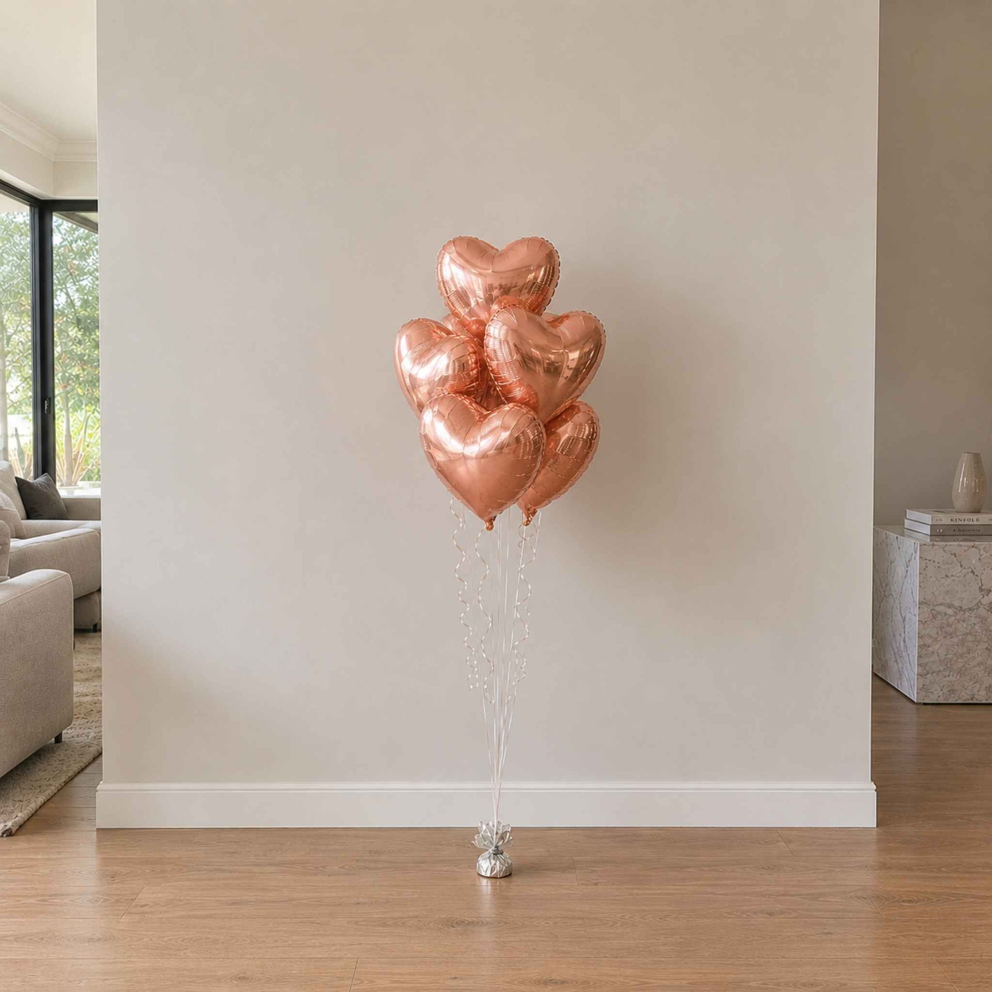 Rose gold heart-shaped balloons on a stand against a neutral wall in a living room.