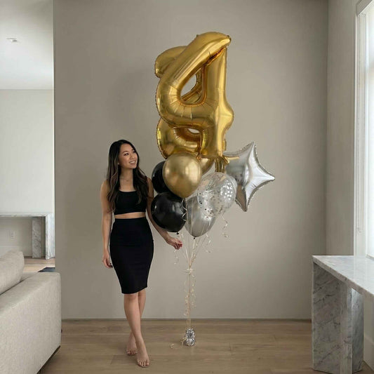 Woman holding a bouquet of gold, black, and silver balloons shaped like numbers in a modern living room.
