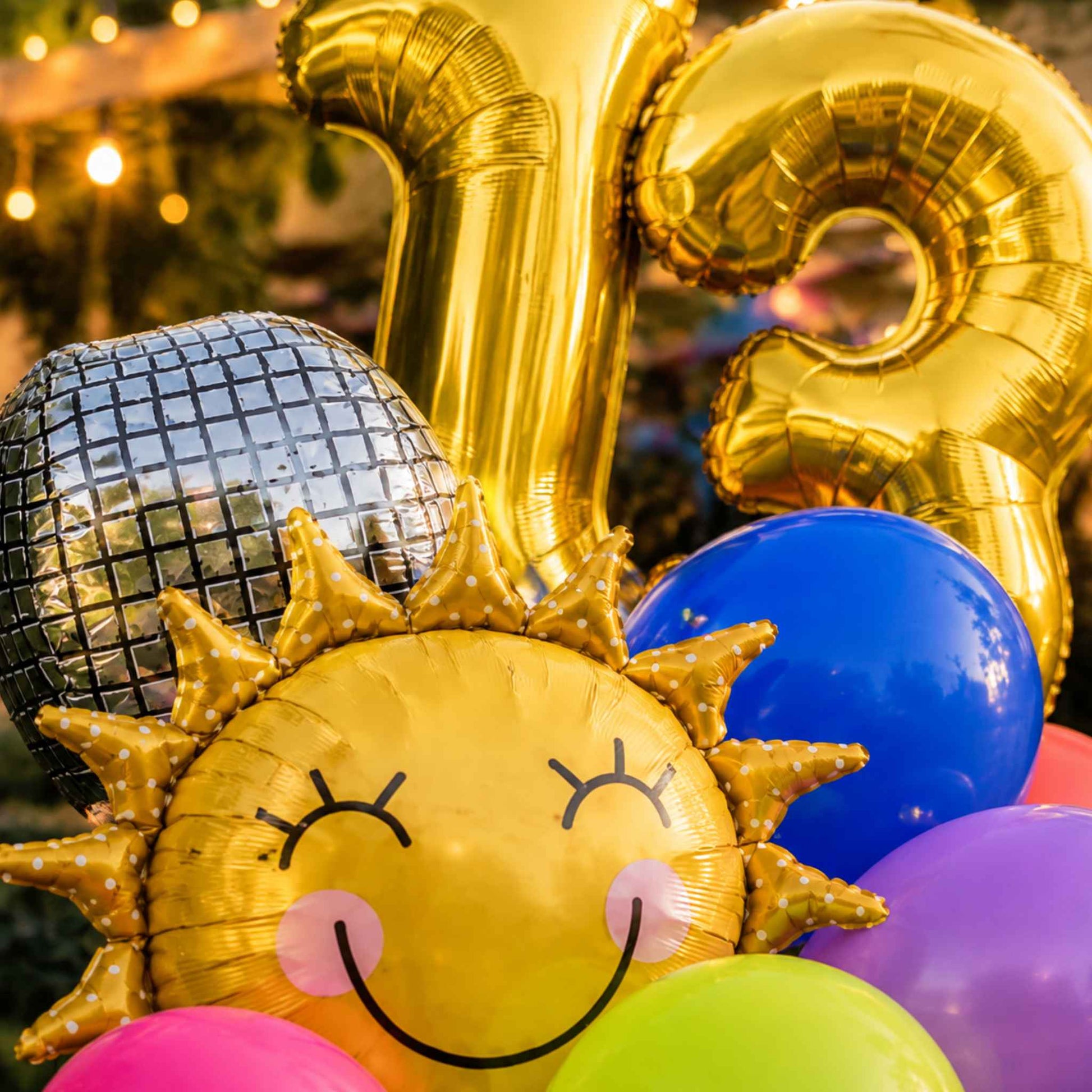A close-up of party balloons including a large gold foil number 13 balloon, a smiling sun face balloon, a disco ball balloon, and several colorful round balloons in blue, purple, pink, and green.