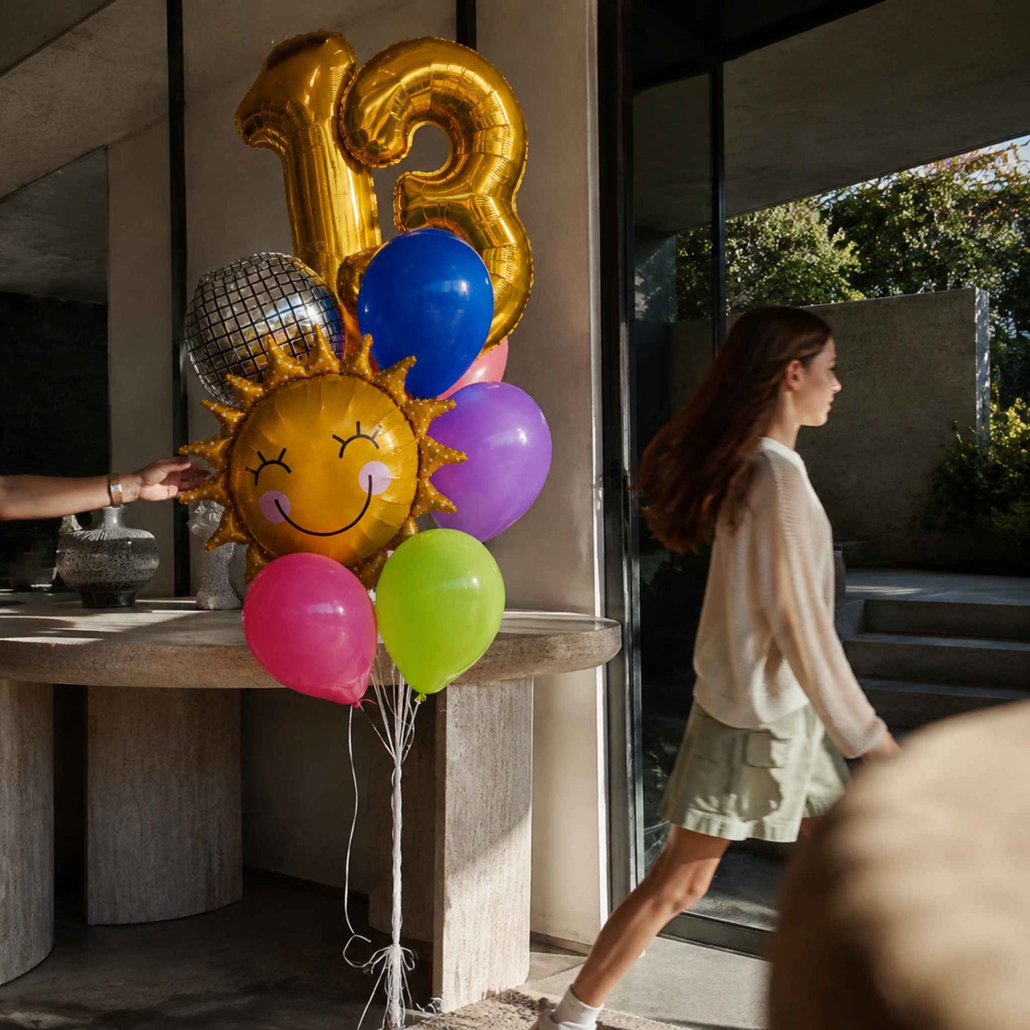 A cluster of foil balloons including gold number 13 balloons, a smiling sun balloon, and colorful round balloons in blue, purple, green, and pink, set next to a woman walking in a modern home interior.