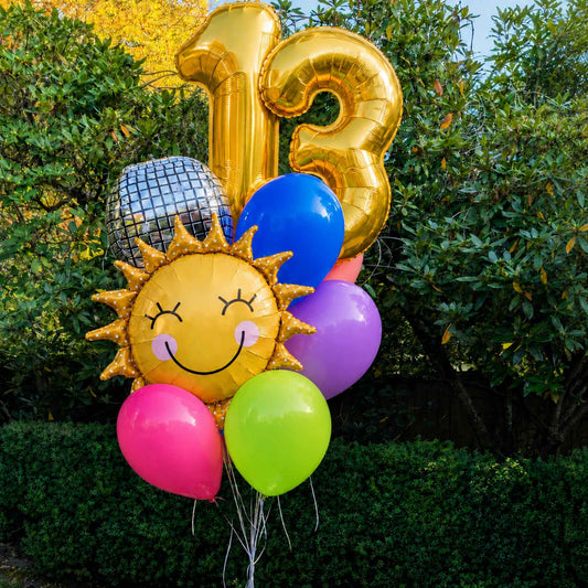 A cluster of party balloons including gold foil number 13 balloons, a smiling sun face balloon, a silver disco ball balloon, and several solid balloons in blue, purple, green, and pink, set against a background of green bushes and trees.