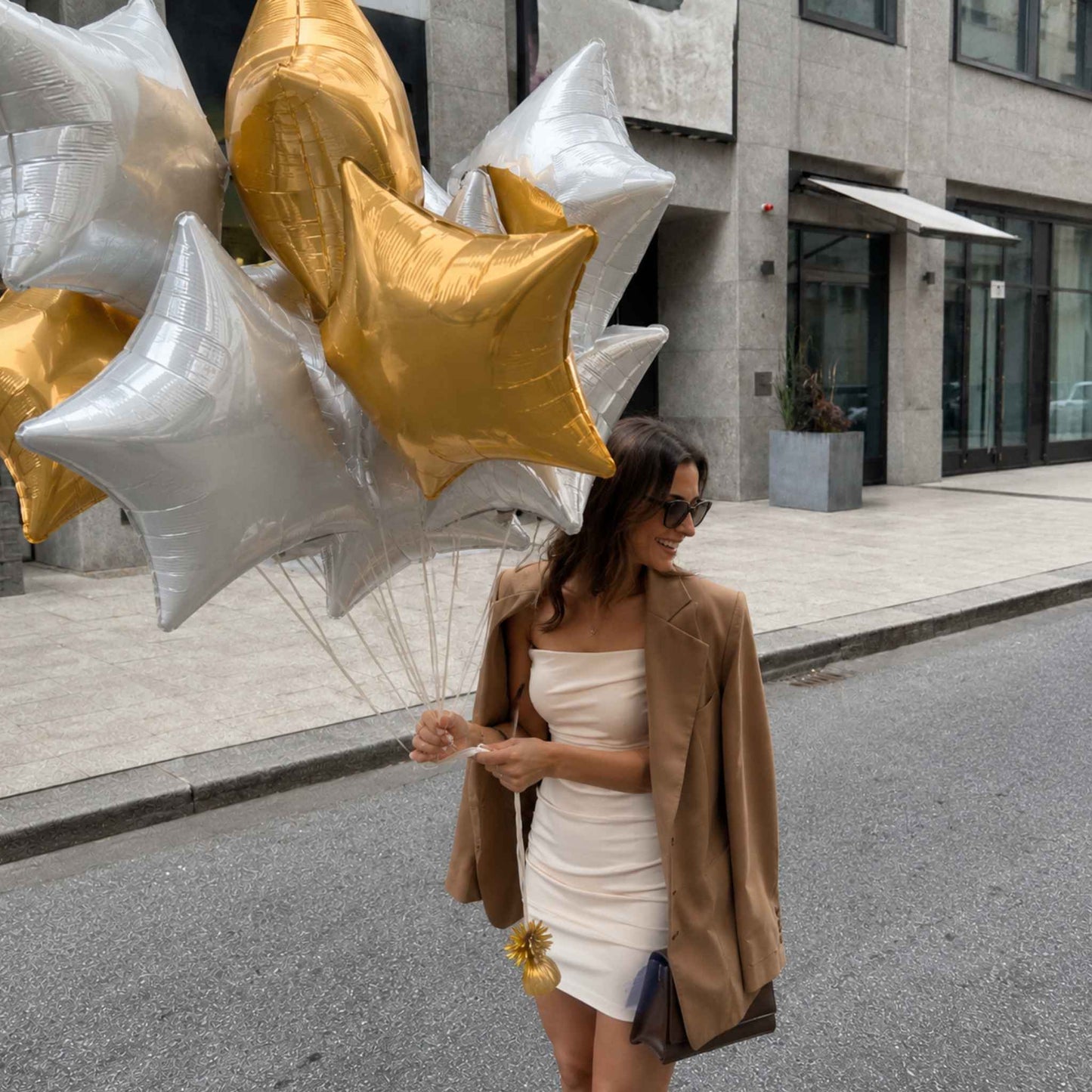 A woman walking on a city street holding a cluster of metallic star-shaped balloons in silver and gold. She is wearing a white strapless mini dress, a tan blazer, and sunglasses.