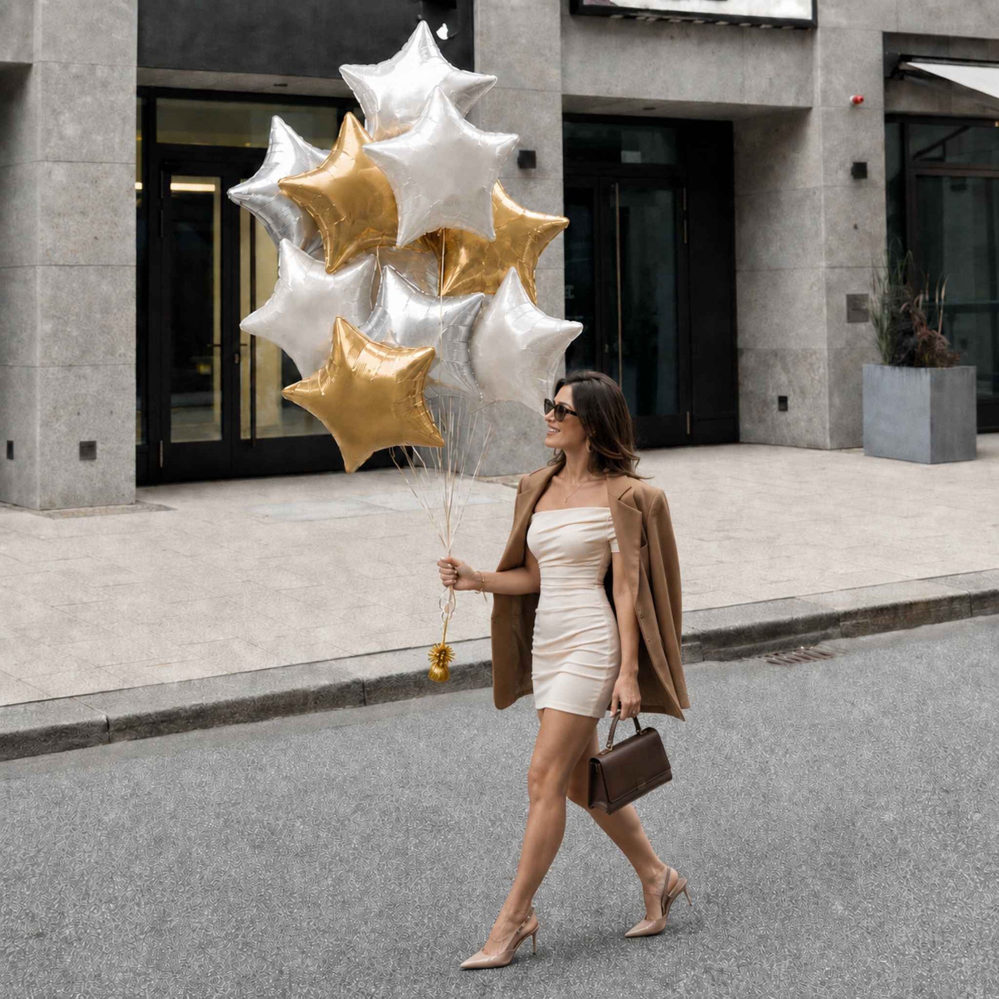 A woman walks on a city street holding a cluster of metallic star-shaped balloons in silver and gold. She is wearing a white strapless mini dress, a tan blazer, and beige heels, and carries a brown handbag.
