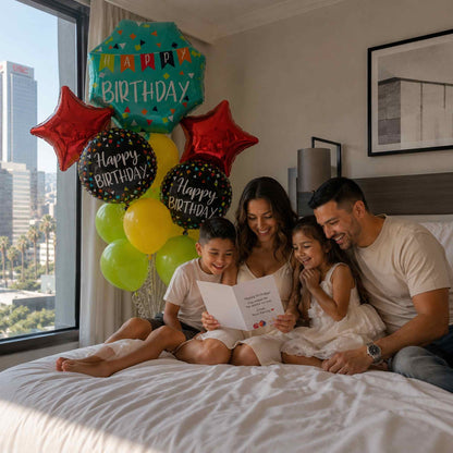 A family of four sits on a bed in a bedroom, reading a birthday card. Large colorful 'Happy Birthday' balloons are clustered near the window on the left, and the room has a city view outside.