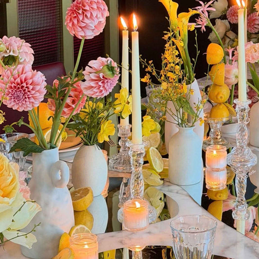 A styled table centerpiece with white ceramic vases filled with pink and yellow flowers, clear glass candlesticks with lit taper candles, and sliced lemons on a reflective surface.
