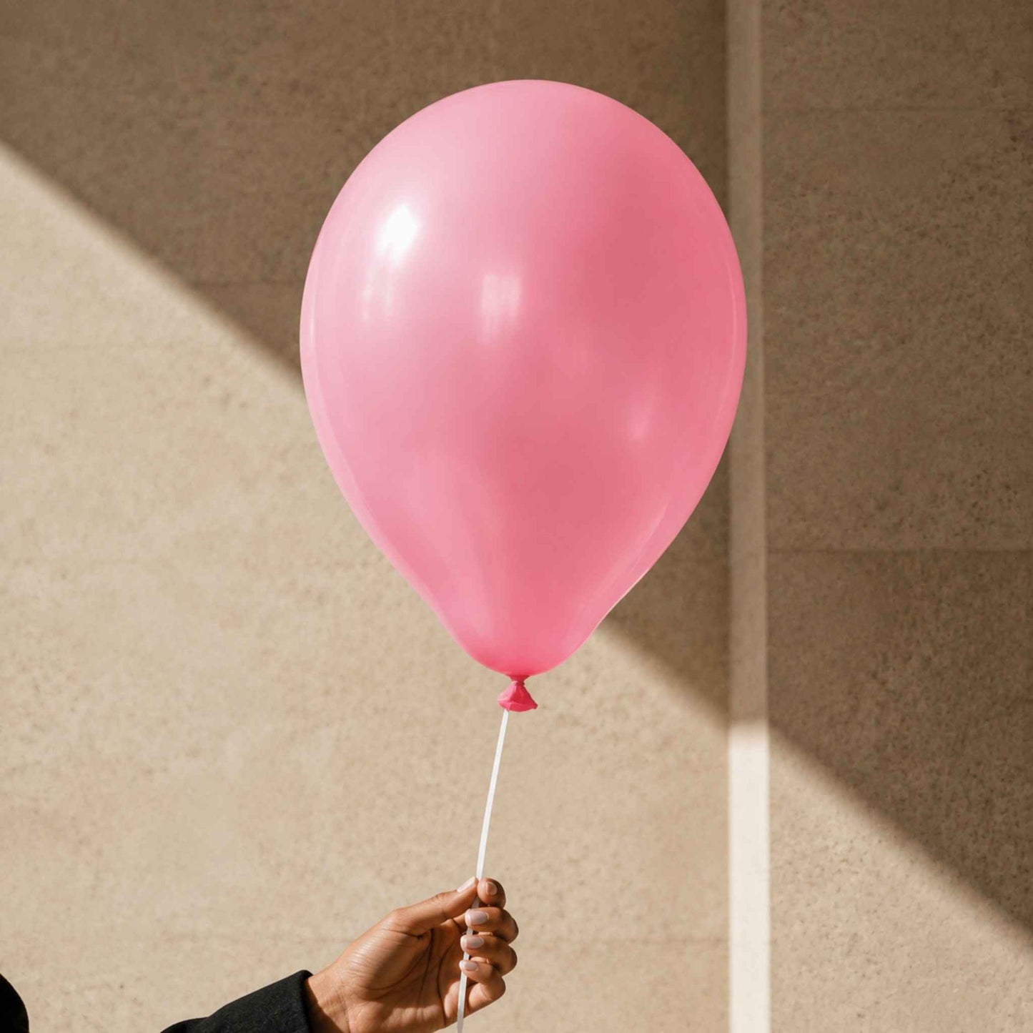 A single matte pink balloon held by a hand against a warm beige textured wall with diagonal sunlight and shadow.