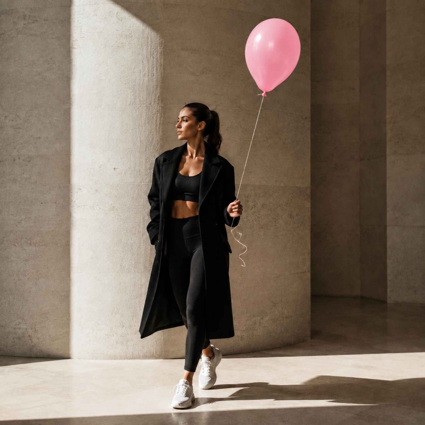 A woman in a black activewear set and long black coat walks through a modern concrete corridor while holding a pink balloon.