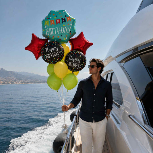 A man wearing sunglasses, a dark navy button-up shirt, and white trousers stands on a white yacht holding a colorful 'Happy Birthday' balloon bouquet against a background of blue sea and sky.
