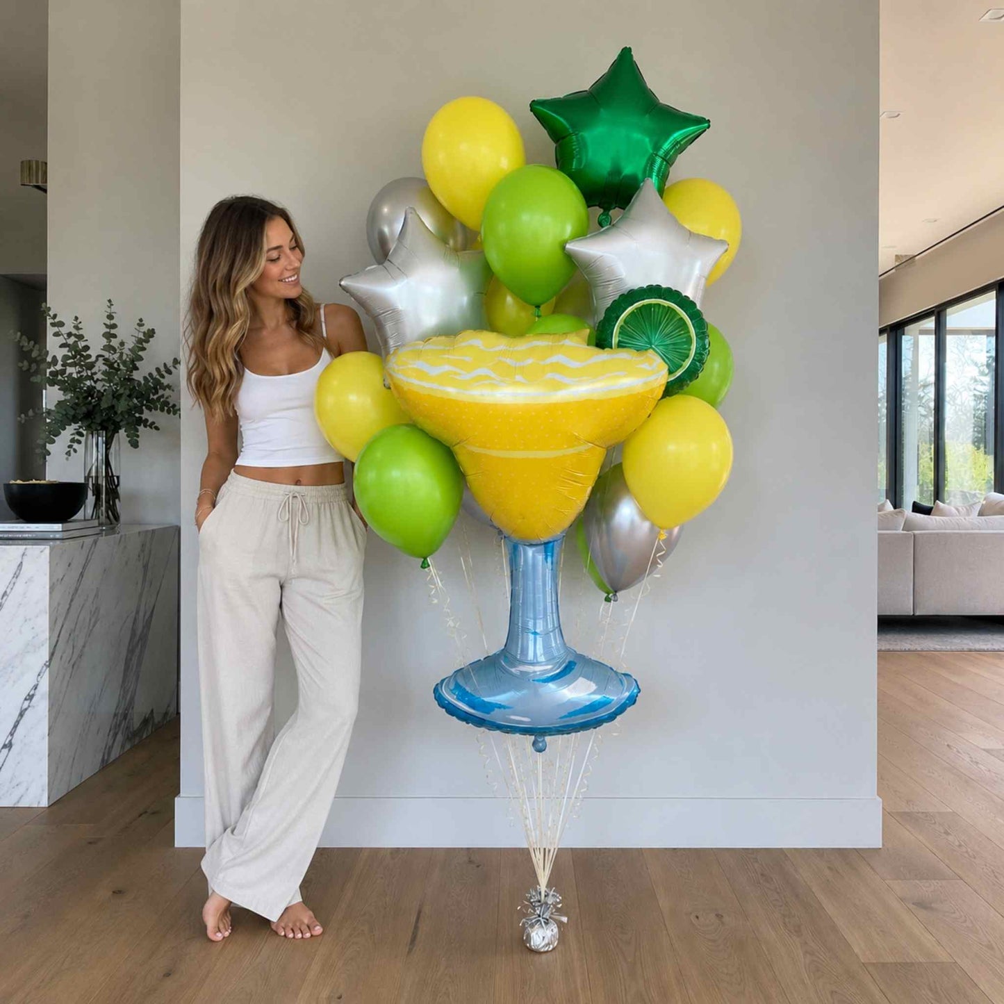 Woman standing next to a large arrangement of colorful balloons, including a large margarita glass balloon, in a modern living room.