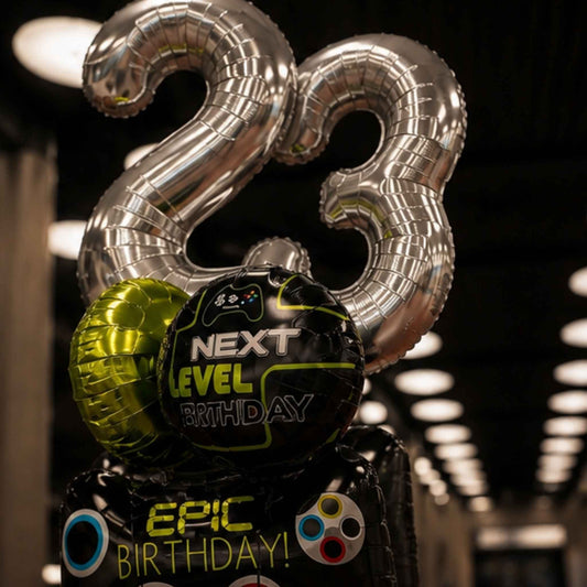A cluster of birthday balloons, including two large metallic silver number 23 balloons, a black round balloon reading 'NEXT LEVEL BIRTHDAY' with a game controller graphic, and a black balloon reading 'EPIC BIRTHDAY!' with colorful controller-style icons, photographed in a dim indoor setting with blurred lights in the background.