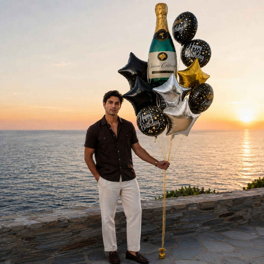 A man in a dark brown short-sleeve button-up shirt and white pants stands on a stone ledge overlooking the ocean at sunset, holding a balloon bouquet styled like a champagne bottle with black, silver, and gold foil balloons.