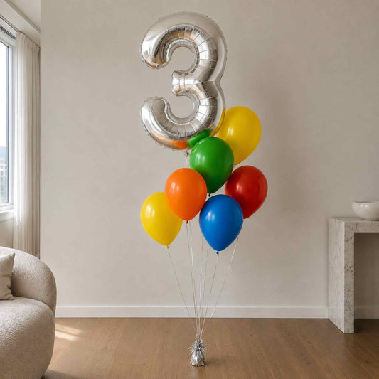 A birthday balloon bouquet with a large metallic silver number 3 foil balloon and six colorful latex balloons in yellow, green, orange, red, and blue, standing on a small silver base in a bright room.