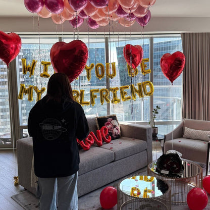 Person standing in a room with heart-shaped balloons and 'Will You Be My Girlfriend?' message.