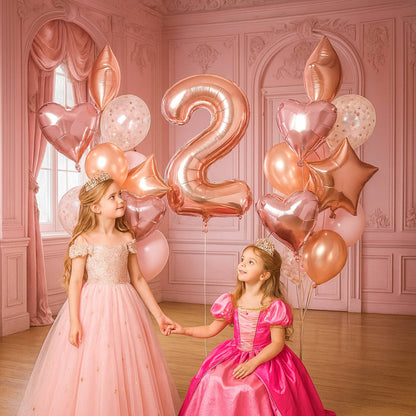 Two young girls in pink dresses holding hands with rose gold balloons shaped like numbers and stars in a decorated room.