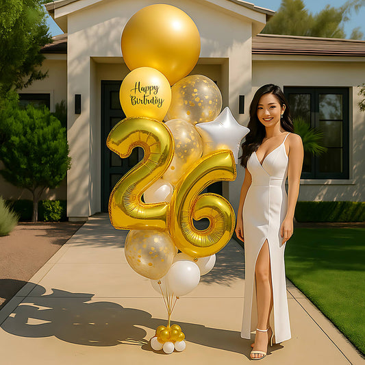 Woman in a white dress standing next to a large 'Happy Birthday' balloon bouquet with number '26' in front of a house.