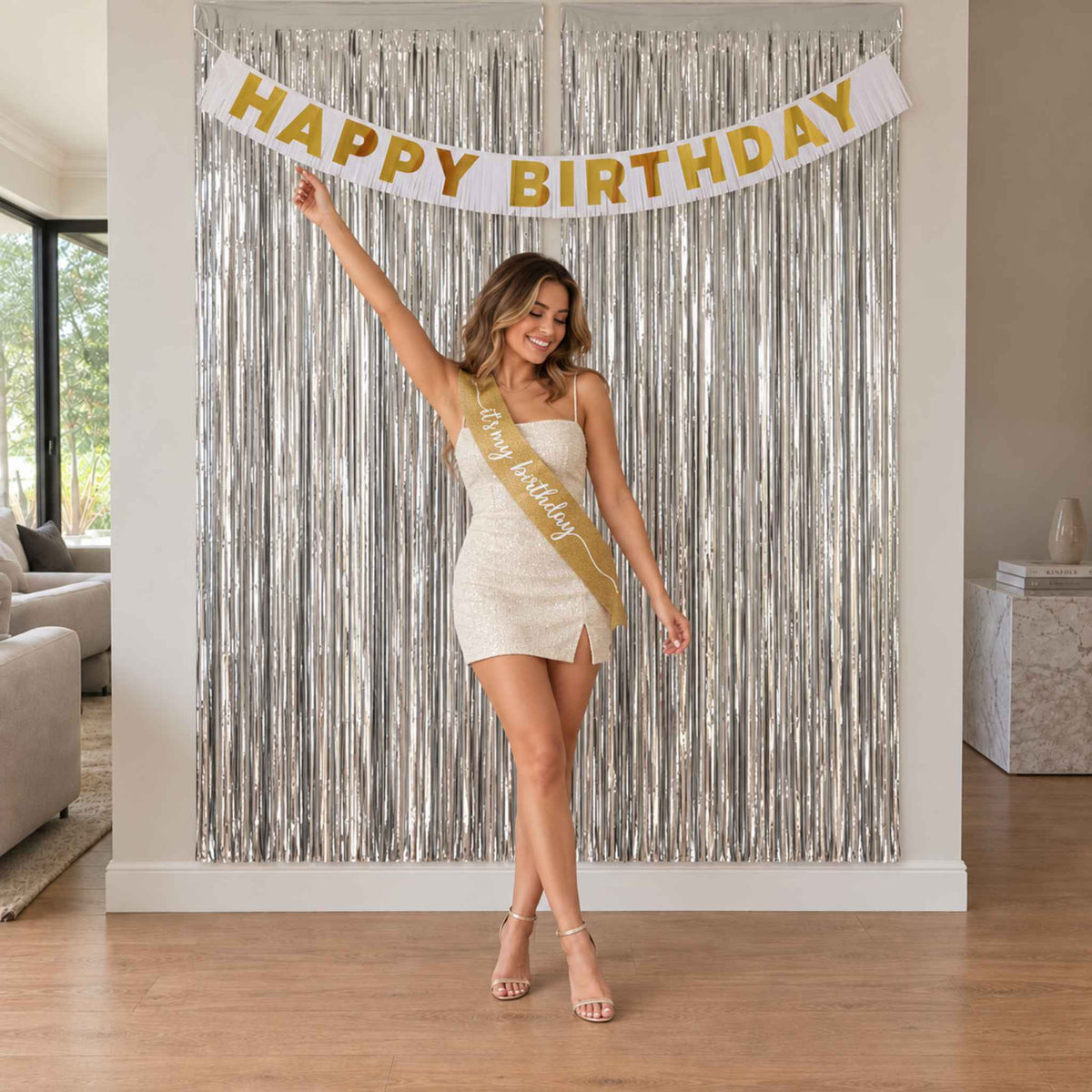A woman stands in a living room in front of a silver tinsel curtain backdrop, holding a white 'HAPPY BIRTHDAY' banner above her and wearing a gold sash that reads 'Happy Birthday.'.