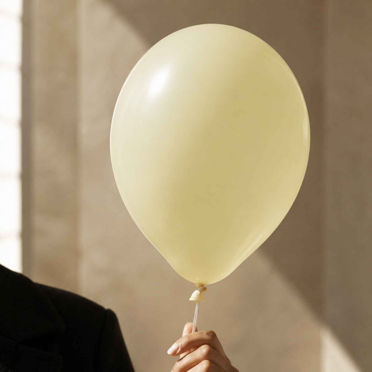 A pale yellow latex balloon held by a person wearing a black blazer, with warm sunlight casting shadows on a beige wall background.