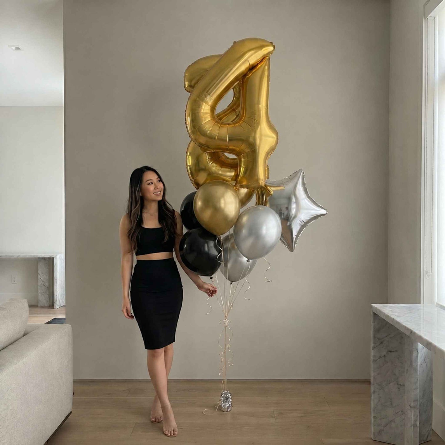 A woman in a black two-piece outfit stands indoors next to a cluster of foil balloons, including a large gold balloon shaped like the number 4, a silver star balloon, and black and silver round balloons.