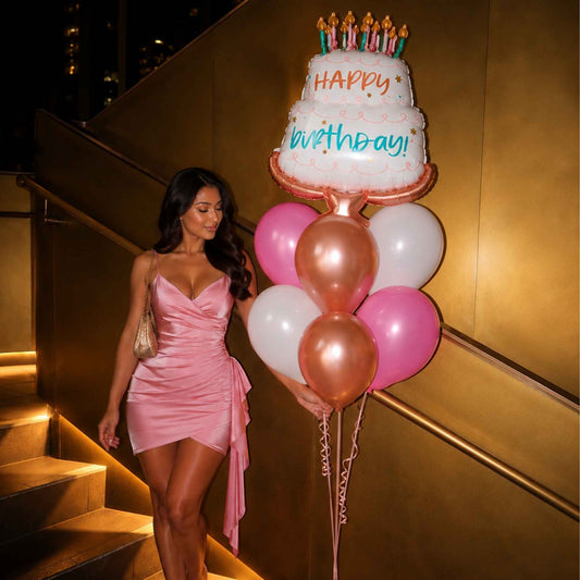 A woman in a pink dress stands on illuminated stairs at night next to a large birthday balloon shaped like a two-tier cake with candles and a cluster of pink, rose gold, and white balloons. The cake balloon reads 'Happy Birthday!'.
