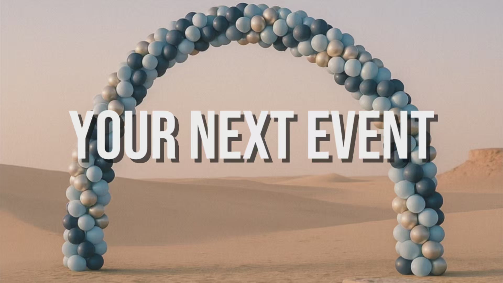 Load video: Large balloon arch install with blue light blue and silver balloons in a dessert setting with soft sand and a base plate