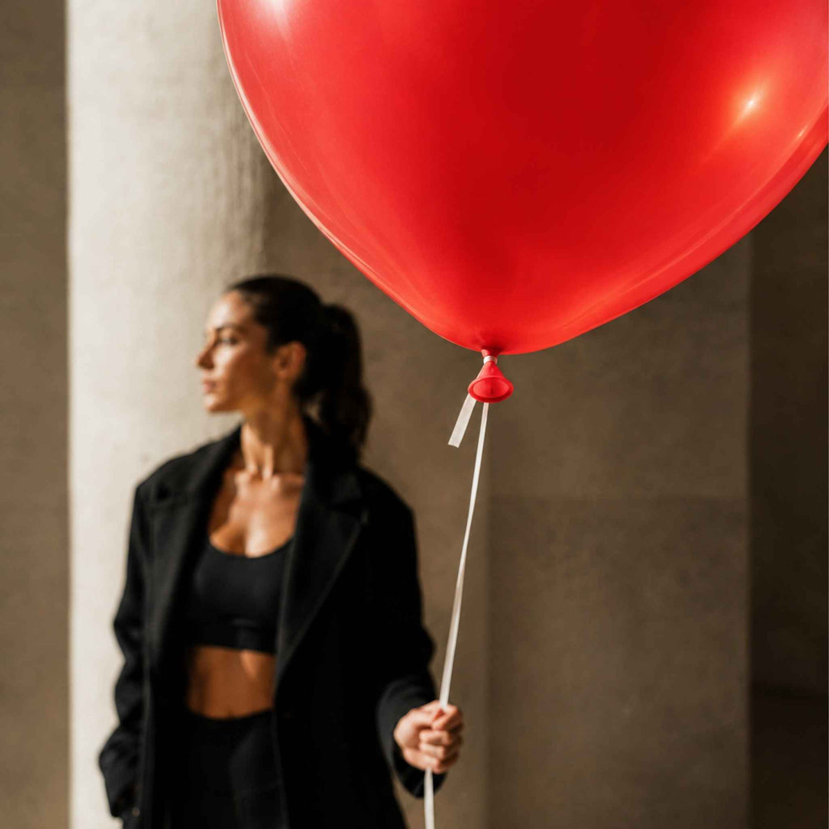 A woman in a black blazer and black sports bra stands in a blurred background while holding the string of a large red balloon that dominates the foreground.