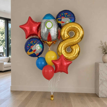 A cluster of metallic and printed birthday balloons arranged indoors against a beige wall, including a large gold number 8 balloon, red star balloons, a blue rocket-shaped balloon, and other space-themed round balloons.