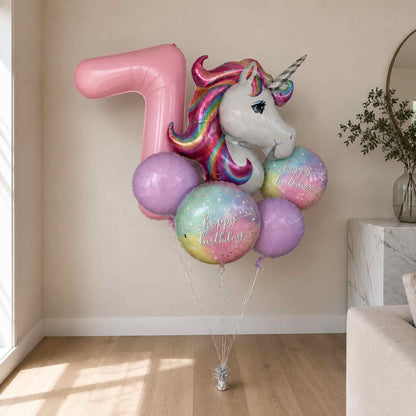 A birthday balloon bouquet featuring a large pink number 7 balloon, a white unicorn head balloon with a rainbow mane, and several pastel round balloons with 'Happy Birthday' text, displayed indoors against a beige wall.