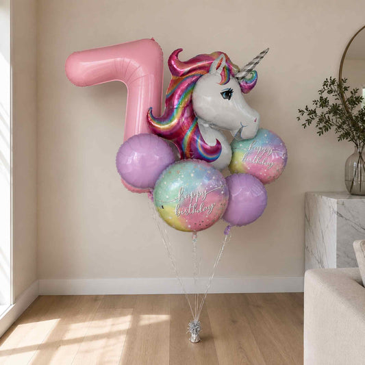 A birthday balloon bouquet featuring a large pink number 7 balloon, a white unicorn head balloon with a rainbow mane, and several pastel round balloons with 'Happy Birthday' text, displayed indoors against a beige wall.